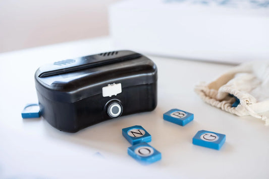 Br’eye braille learning device on a table with blue letter tiles and a drawstring bag beside it